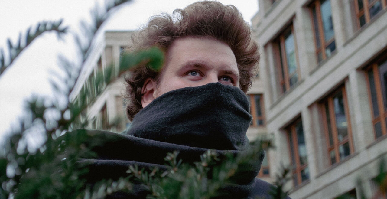 Rainer Winkler Drachenlord - Zu sehen ist der YouTuber und Streamer Rainer Winkler alias „Drachenlord“. Er hat braune Haare und trägt einen schwarzen Hoodie. Über dem Mund hat er einen schwarzen Schal drapiert. Er steht vor einem braunbeigen Bürohaus, im Vordergrund sind die Äste eines Nadelbaums zu sehen, hinter dem er sich zu verstecken scheint. - © Foto: IMAGO / Pond5 Images
