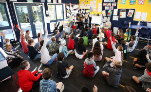Eine Volksschule in Auckland, Neuseeland - Diese Aufnahme von schräg oben zeigt ein Klassenzimmer voller Schülerinnen und Schüler, die am Boden sitzen und einer Lehrkraft zuhören.  - © Getty Images / Fiona Goodall