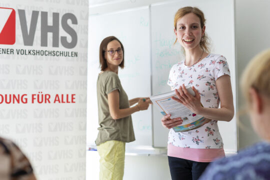 Basisbildung und Alphabetisierung an der Volkshochschule Wien - Man sieht eine Lehrerin und mehrere erwachsene Teilnehmende in einem Kurs für Basisbildung. Im Hintergrund ist das Logo der Volkshochschule zu sehen.  - © VHS Favoriten