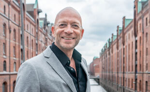 Eric Hegmann, Paartherapeut - Das Porträt zeigt Eric Hegmann in einem grauen Anzug und schwarzem Hemd in der Speicherstatt in Hamburg.  - © Robert Hilton
