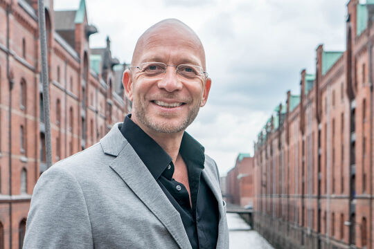 Eric Hegmann, Paartherapeut - Das Porträt zeigt Eric Hegmann in einem grauen Anzug und schwarzem Hemd in der Speicherstatt in Hamburg.  - © Robert Hilton