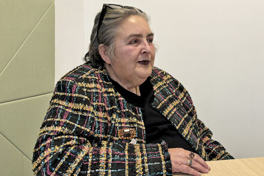 Eva Blimlinger - Eva Blimlinger sitzt an einem Tisch, hinter ihr sind eine weiße Wand und eine hellgrüne Wand zu sehen. Sie hat den rechten Arm am Tisch abgestützt und blickt nach links. Sie trägt eine bunt gemusterte Jacke und eine schwarze Bluse. In ihren Haaren steckt eine schwarze Brille. - © Foto: Viktoria Kapp