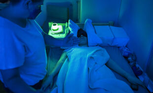 ME/CFS Patient - Person liegt mit Schlafmaske und Decke auf einer Liege, daneben steht eine weitere Liege und eine Person hält die Hand der liegenden Person. - © Foto: We&Me Foundation / Brent Stirton