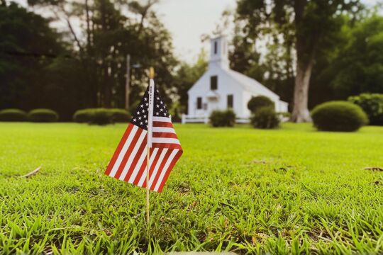 Flagge USA Kirche - Eine kleine Flagge der USA steckt in einer grünen Wiese vor einer Kirche. - © Pexels / Shelby Cox