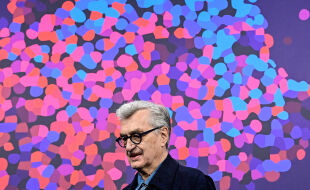Wim Wenders Berlinale - Wim Wenders trägt einen dunklen Mantel vor einem Hintergrund aus bunten Flecken, die einen Bären stilisieren. - © APA / AFP / John MacDougall