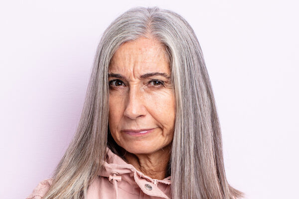 Wütende ältere Frau - Das Foto zeigt eine ältere Frau mit glatten, grauen Haaren und wütendem Gesichtsausdruck inklusive Zornesfalte vor einem hellrosa Hintergrund. - © Adobe Stock / kues1