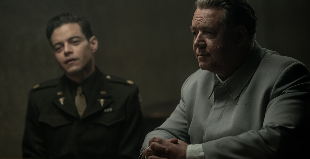 Nürnberg Film - Szenenbild aus dem Film „Nürnberg“ mit Russell Crowe (rechts) und Rami Malek (links): Im Vordergrund rechts sitzt ein älterer Mann mit grauen Haaren an einem Tisch, seine Hände liegen ineinander verschränkt auf der Tischplatte. Er trägt eine hellgraue einfache Uniformjacke, unter dem Ärmel ist ein weißes Hemd zu sehen. Im Hintergrund ist leicht verschwommen ein jüngerer Mann mit schwarzen Haaren zu sehen. Er trägt eine US-amerikanische dunkelbraune Militäruniform und eine beige Krawatte. - © Foto: Polyfilm