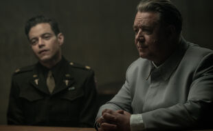 Nürnberg Film - Szenenbild aus dem Film „Nürnberg“ mit Russell Crowe (rechts) und Rami Malek (links): Im Vordergrund rechts sitzt ein älterer Mann mit grauen Haaren an einem Tisch, seine Hände liegen ineinander verschränkt auf der Tischplatte. Er trägt eine hellgraue einfache Uniformjacke, unter dem Ärmel ist ein weißes Hemd zu sehen. Im Hintergrund ist leicht verschwommen ein jüngerer Mann mit schwarzen Haaren zu sehen. Er trägt eine US-amerikanische dunkelbraune Militäruniform und eine beige Krawatte. - © Foto: Polyfilm