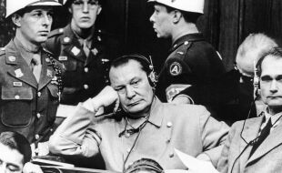 Hermann Göring Nürnberger Prozesse - Schwarz-weiß-Foto von Hermann Göring vor Gericht in Nürnberg. Er sitzt auf der Anklagebank und trägt eine hellgraue doppelreihige Uniformjacke. Er trägt Kopfhörer mit Kabel und stützt seinen Kopf auf seiner rechten Hand ab. Rechts neben ihm ist sein Mitangeklagter Rudolf Heß zu sehen. Um sie herum sind drei Wachebeamte in Uniform und mit weißen Helmen zu sehen. - © Foto: IMAGO / ITAR-TASS