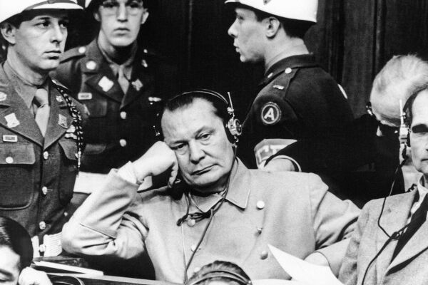 Hermann Göring Nürnberger Prozesse - Schwarz-weiß-Foto von Hermann Göring vor Gericht in Nürnberg. Er sitzt auf der Anklagebank und trägt eine hellgraue doppelreihige Uniformjacke. Er trägt Kopfhörer mit Kabel und stützt seinen Kopf auf seiner rechten Hand ab. Rechts neben ihm ist sein Mitangeklagter Rudolf Heß zu sehen. Um sie herum sind drei Wachebeamte in Uniform und mit weißen Helmen zu sehen. - © Foto: IMAGO / ITAR-TASS