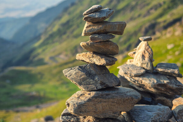Steinhaufen Meditation Kontemplation Stille - Mehrere unregelmäßig geformte Steine sind zu zwei kleinen Steinhaufen aufeinander gestapelt, im Hintergrund unscharfe grüne Hügel und Berge - © Foto: Adobe Stock / Daniel CHETRONI