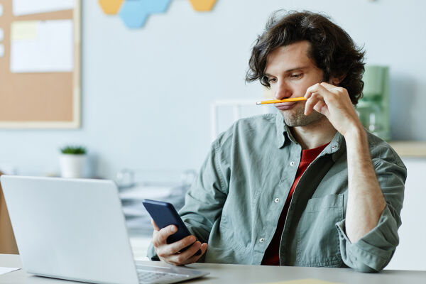 Prokrastination - Das Foto zeigt einen jungen Mann in seinem Büro, der missmutig auf sein Handy starrt. - © Adobe Stock /  Seventyfour