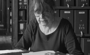 Arlette Farge - Arlette Farge mit hochgestecktem Haar und schwarzem Oberteil sitzt an einem Tisch vor einem geöffneten Buch in einem Raum mit Regalen voller beschrifteter Ordner. - © Foto: Wikipedia/REMOLOC (CC BY-SA 4.0)
