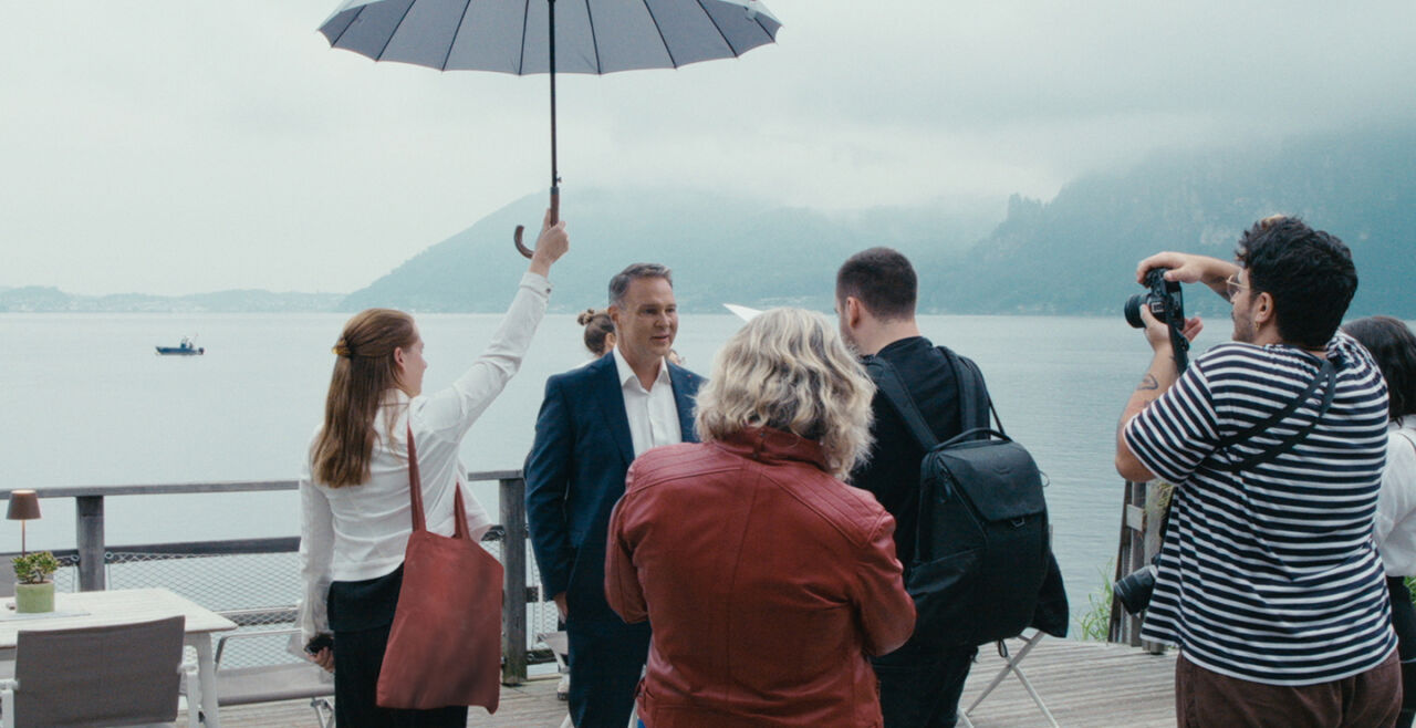 Wahlkampf - Personen auf einer Terrasse am Wasser, eine Frau hält einen Regenschirm über einen Mann im Anzug, ein Fotograf macht ein Foto - © Filmladen