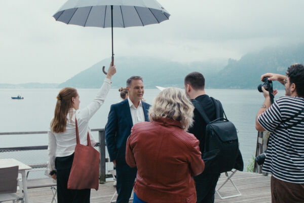 Wahlkampf - Personen auf einer Terrasse am Wasser, eine Frau hält einen Regenschirm über einen Mann im Anzug, ein Fotograf macht ein Foto - © Filmladen