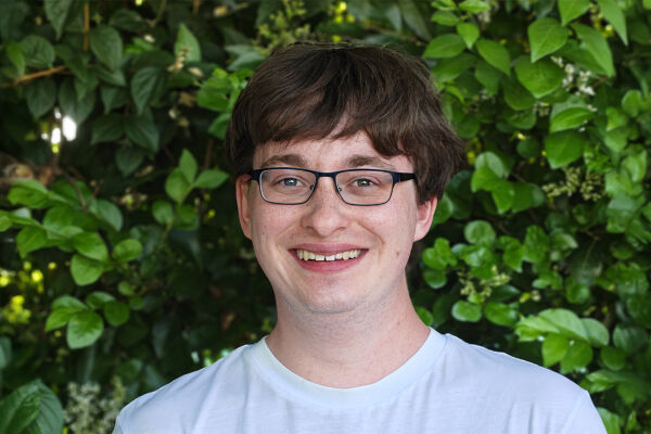 Felix Zych - Porträtfoto von Felix Zych. Er hat kurze braune Haare und trägt eine schwarze Brille und ein weißes T-Shirt. Er lächelt in die Kamera. Im Hintergrund ist eine grüne Hecke zu sehen. - © Foto: Privat