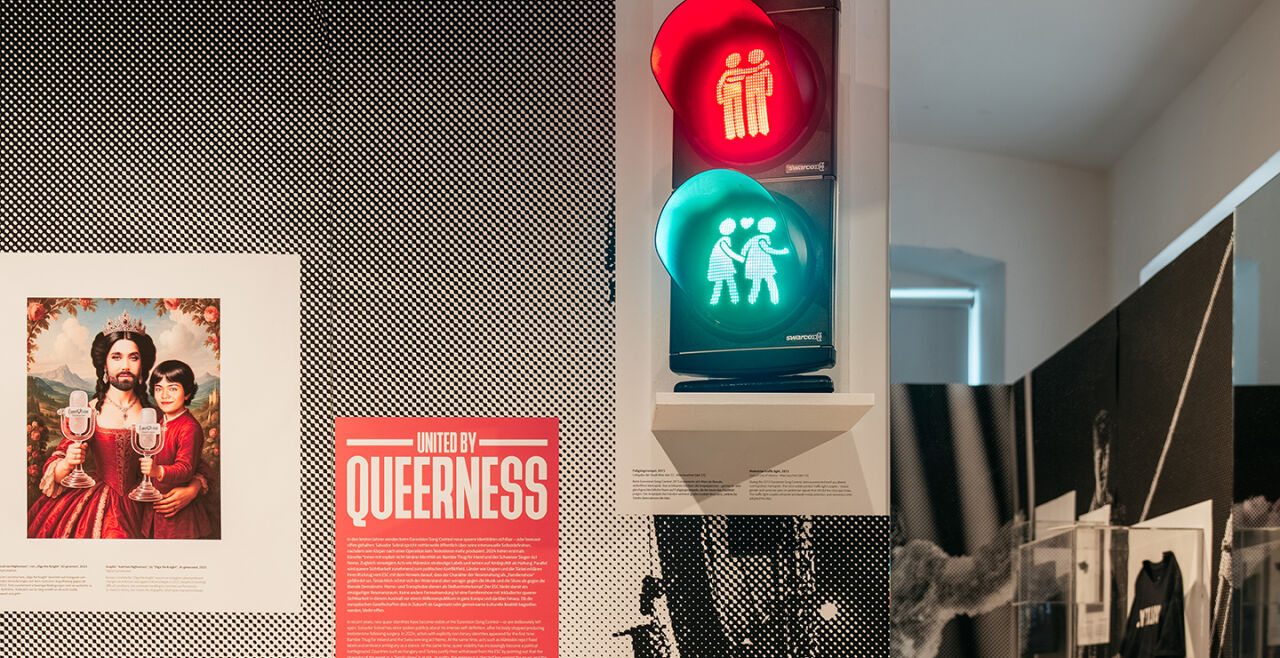 Ausstellung "United by Queerness" im Qwien - Foto aus der Ausstellung "United by Queerness" im Qwien. Zu sehen ist eine Infotafel mit dem Ausstellungstitel „United by Queerness“. In der Mitte ist eine Ampel zu sehen, auf der sowohl rot als auch grün leuchtet. Statt einzelnen Ampelmännchen sind darin die Wiener Ampelpärchen zu sehen, oben in rotem Licht zwei Männer, unten im grünen Licht zwei Frauen. Rechts davon ist ein Spiegel zu sehen. Links davon ist eine Grafik zu sehen, in der die Song-Contest-Siegerin Conchita in einem Königinnen-ähnlichen roten Kleid dargestellt ist. In der rechten Hand hält sie den Eurovision-Pokal, auf dem linken Arm hält sie eine jungen Mann, der dem Musiker JJ ähnelt, der 2025 den Song Contest ebenfalls für Österreich gewann. Auch er hält eine Eurovision-Trophäe. - © Foto: Gregor Hofbauer