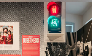 Ausstellung "United by Queerness" im Qwien - Foto aus der Ausstellung "United by Queerness" im Qwien. Zu sehen ist eine Infotafel mit dem Ausstellungstitel „United by Queerness“. In der Mitte ist eine Ampel zu sehen, auf der sowohl rot als auch grün leuchtet. Statt einzelnen Ampelmännchen sind darin die Wiener Ampelpärchen zu sehen, oben in rotem Licht zwei Männer, unten im grünen Licht zwei Frauen. Rechts davon ist ein Spiegel zu sehen. Links davon ist eine Grafik zu sehen, in der die Song-Contest-Siegerin Conchita in einem Königinnen-ähnlichen roten Kleid dargestellt ist. In der rechten Hand hält sie den Eurovision-Pokal, auf dem linken Arm hält sie eine jungen Mann, der dem Musiker JJ ähnelt, der 2025 den Song Contest ebenfalls für Österreich gewann. Auch er hält eine Eurovision-Trophäe. - © Foto: Gregor Hofbauer