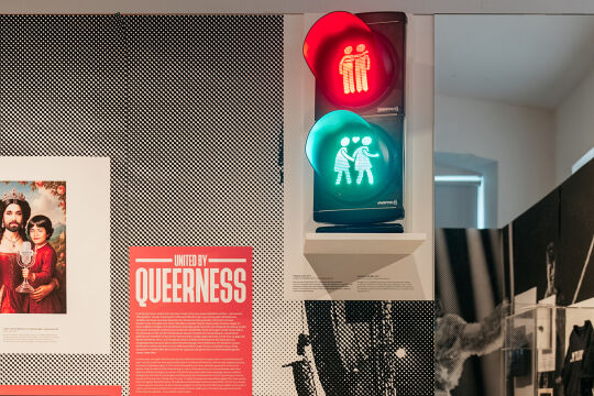 Ausstellung "United by Queerness" im Qwien - Foto aus der Ausstellung "United by Queerness" im Qwien. Zu sehen ist eine Infotafel mit dem Ausstellungstitel „United by Queerness“. In der Mitte ist eine Ampel zu sehen, auf der sowohl rot als auch grün leuchtet. Statt einzelnen Ampelmännchen sind darin die Wiener Ampelpärchen zu sehen, oben in rotem Licht zwei Männer, unten im grünen Licht zwei Frauen. Rechts davon ist ein Spiegel zu sehen. Links davon ist eine Grafik zu sehen, in der die Song-Contest-Siegerin Conchita in einem Königinnen-ähnlichen roten Kleid dargestellt ist. In der rechten Hand hält sie den Eurovision-Pokal, auf dem linken Arm hält sie eine jungen Mann, der dem Musiker JJ ähnelt, der 2025 den Song Contest ebenfalls für Österreich gewann. Auch er hält eine Eurovision-Trophäe. - © Foto: Gregor Hofbauer