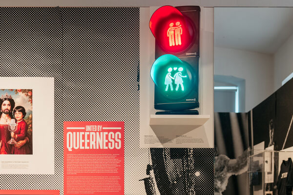 Ausstellung "United by Queerness" im Qwien - Foto aus der Ausstellung "United by Queerness" im Qwien. Zu sehen ist eine Infotafel mit dem Ausstellungstitel „United by Queerness“. In der Mitte ist eine Ampel zu sehen, auf der sowohl rot als auch grün leuchtet. Statt einzelnen Ampelmännchen sind darin die Wiener Ampelpärchen zu sehen, oben in rotem Licht zwei Männer, unten im grünen Licht zwei Frauen. Rechts davon ist ein Spiegel zu sehen. Links davon ist eine Grafik zu sehen, in der die Song-Contest-Siegerin Conchita in einem Königinnen-ähnlichen roten Kleid dargestellt ist. In der rechten Hand hält sie den Eurovision-Pokal, auf dem linken Arm hält sie eine jungen Mann, der dem Musiker JJ ähnelt, der 2025 den Song Contest ebenfalls für Österreich gewann. Auch er hält eine Eurovision-Trophäe. - © Foto: Gregor Hofbauer