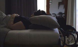 long covid - Kranke Frau im Bett mit Augenmaske in einem verdunkelten Zimmer. Im Hintergrund ist ein Rollstuhl. - © Foto: Lea Aring