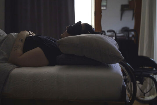 long covid - Kranke Frau im Bett mit Augenmaske in einem verdunkelten Zimmer. Im Hintergrund ist ein Rollstuhl. - © Foto: Lea Aring